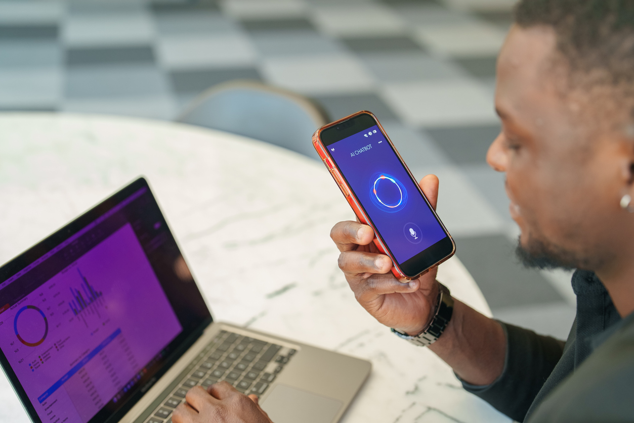 A professional man sitting at a desk, using a voice assistant on his smartphone while analyzing data on his laptop. Concepts of multitasking, modern technology, AI assistants, and digital business solutions.